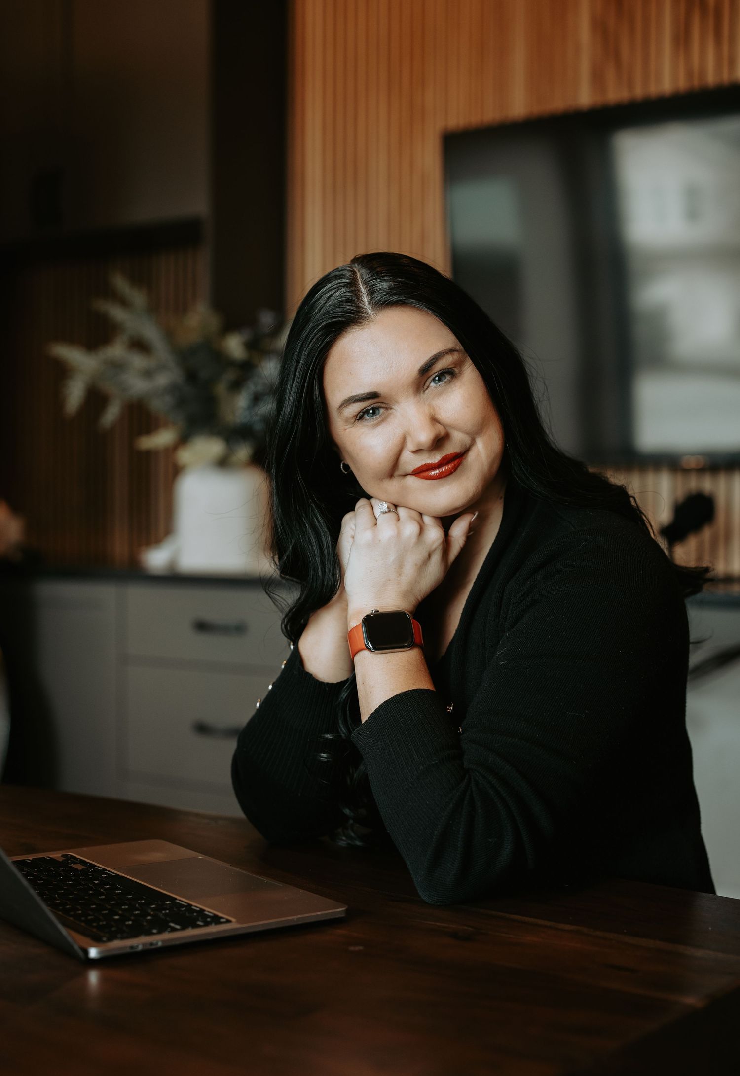 Woman with black hair and red lips smiles at desk with laptop, wearing black top and smartwatch in modern kitchen.