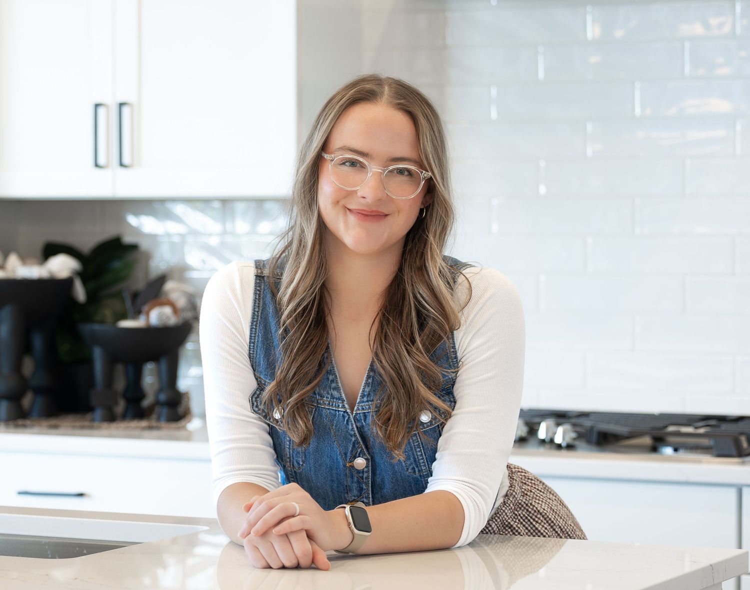 Young woman with long hair and glasses smiling in a modern white kitchen, leaning on a marble countertop.