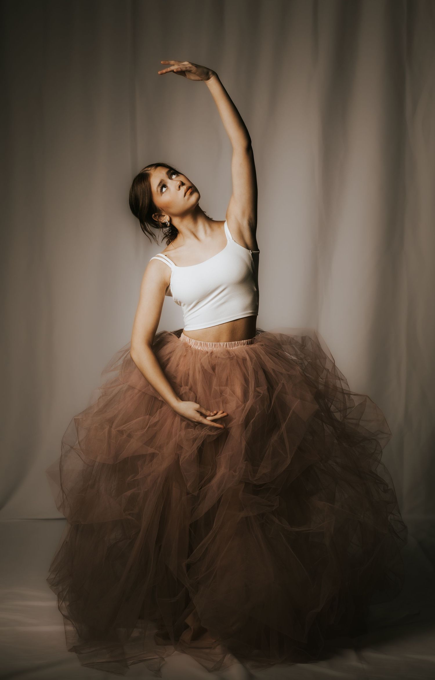 Dancer in white top and flowing brown tulle skirt performs graceful movements in dramatic studio lighting.