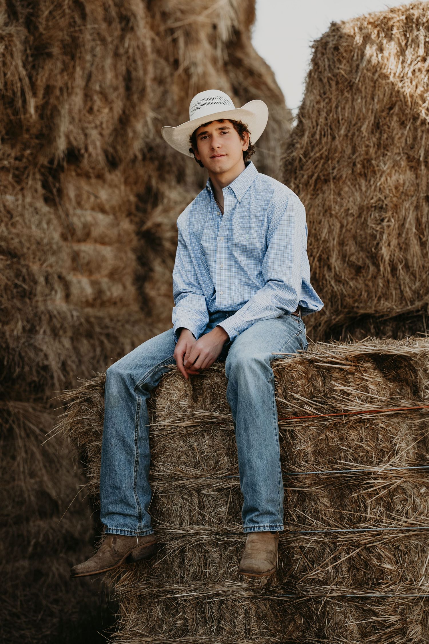 Young man in cowboy hat, plaid shirt, and jeans sitting on hay bales in a rustic outdoor setting.