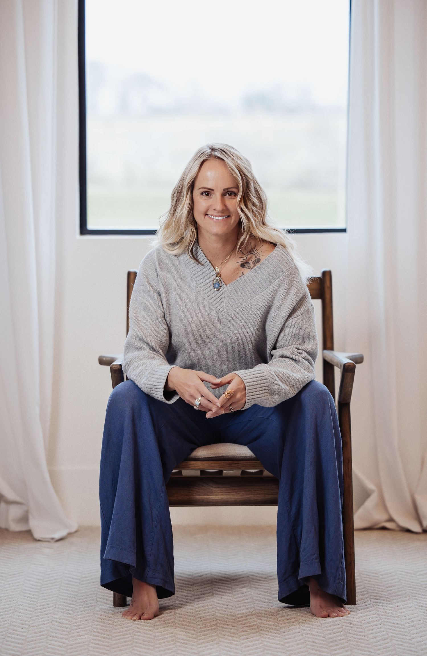Someone sits in a wooden chair wearing a gray sweater and navy wide-leg pants by a window with white curtains.