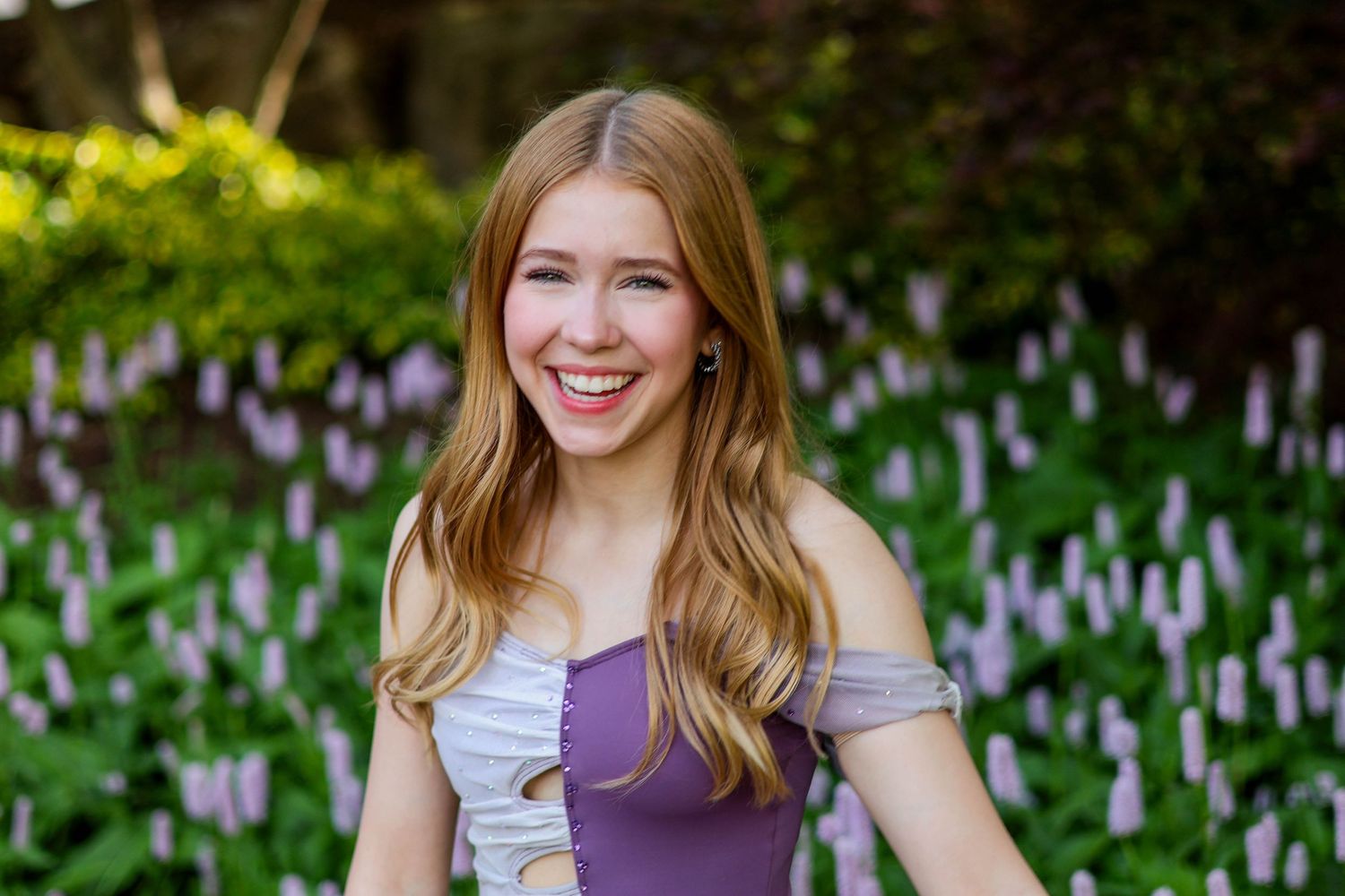 A person in a purple off-shoulder dress poses in a garden filled with white and purple flowers.