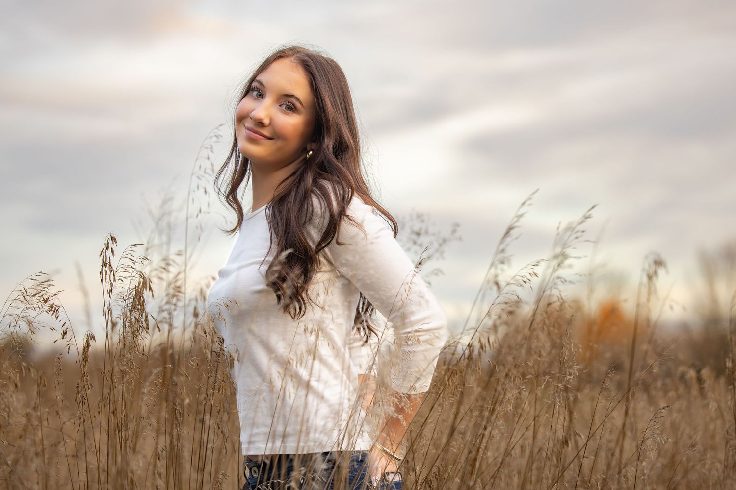 Woman in white sweater poses in tall autumn grass field during golden hour sunset.