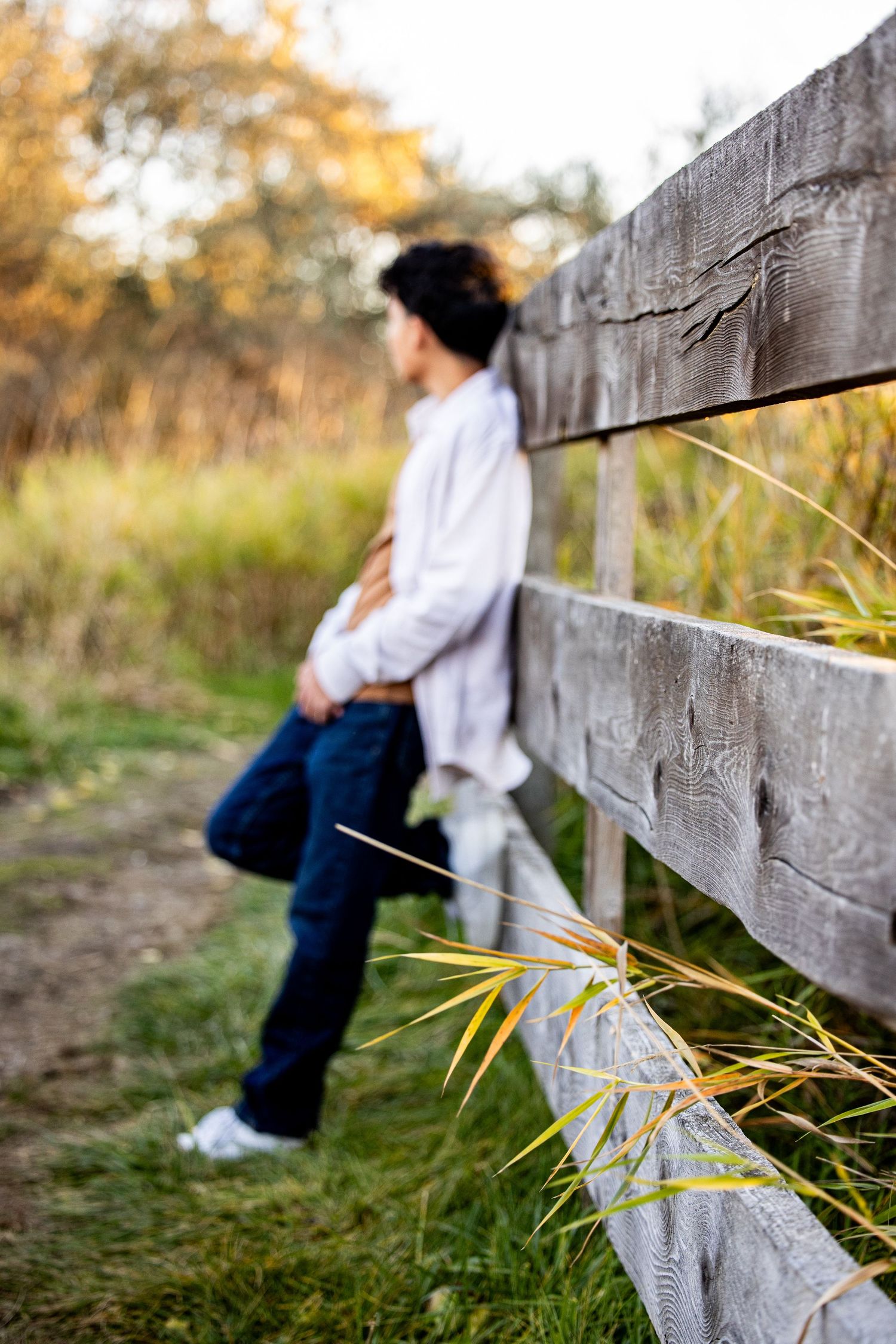 A person in a white shirt and jeans leans against a rustic wooden fence in an autumn field at sunset.