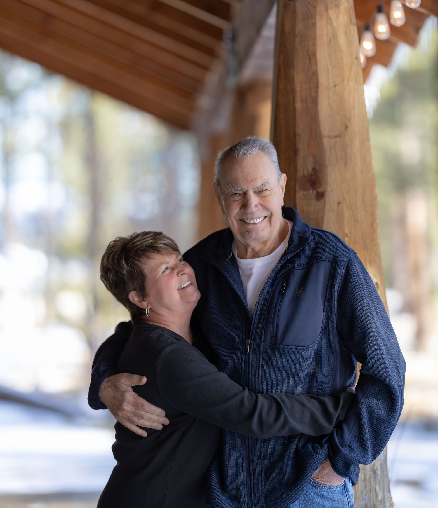 Two people embrace and laugh joyfully under a wooden covered porch area in a candid moment.