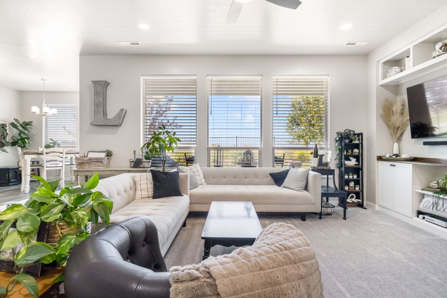 Modern living room with gray carpet, beige sectional sofa, houseplants, and large windows offering natural light.