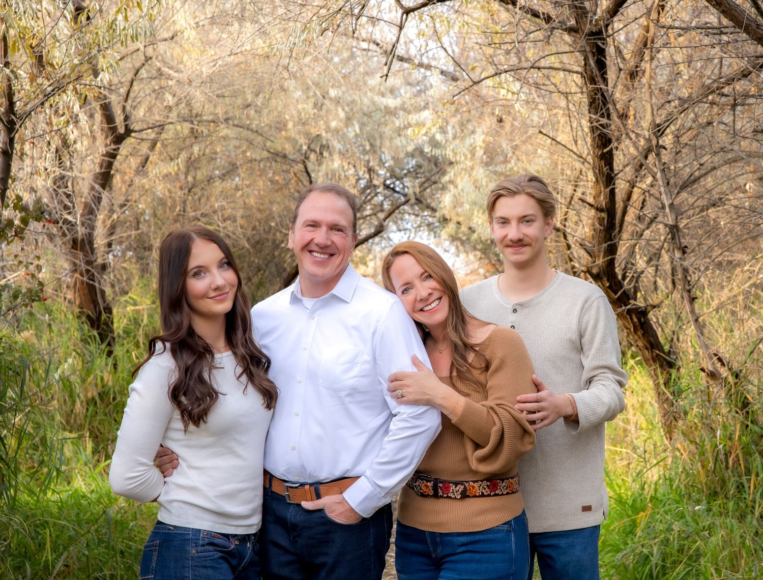 A family of four poses together outdoors in casual attire among autumn trees and tall grass.