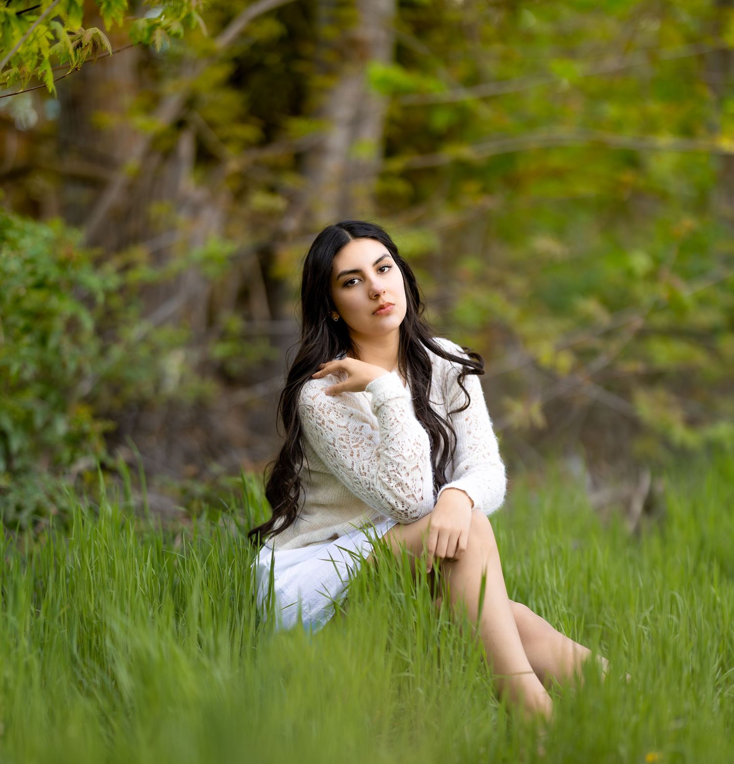Young woman with long dark hair sits in lush green grass wearing a white sweater and skirt, posing outdoors.