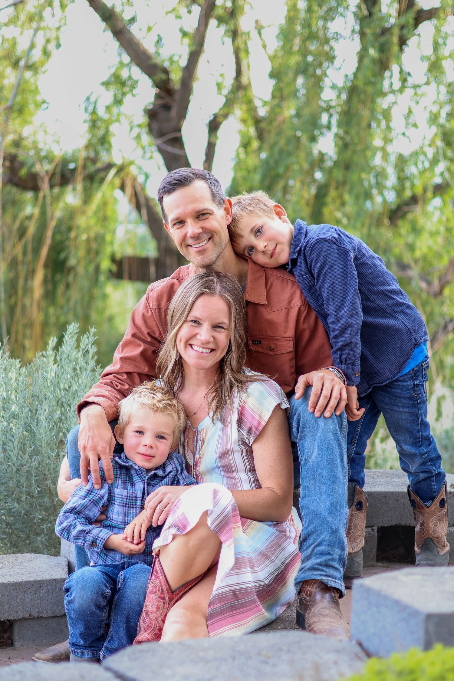 Family of four poses together on stone steps with willow trees and greenery behind them.