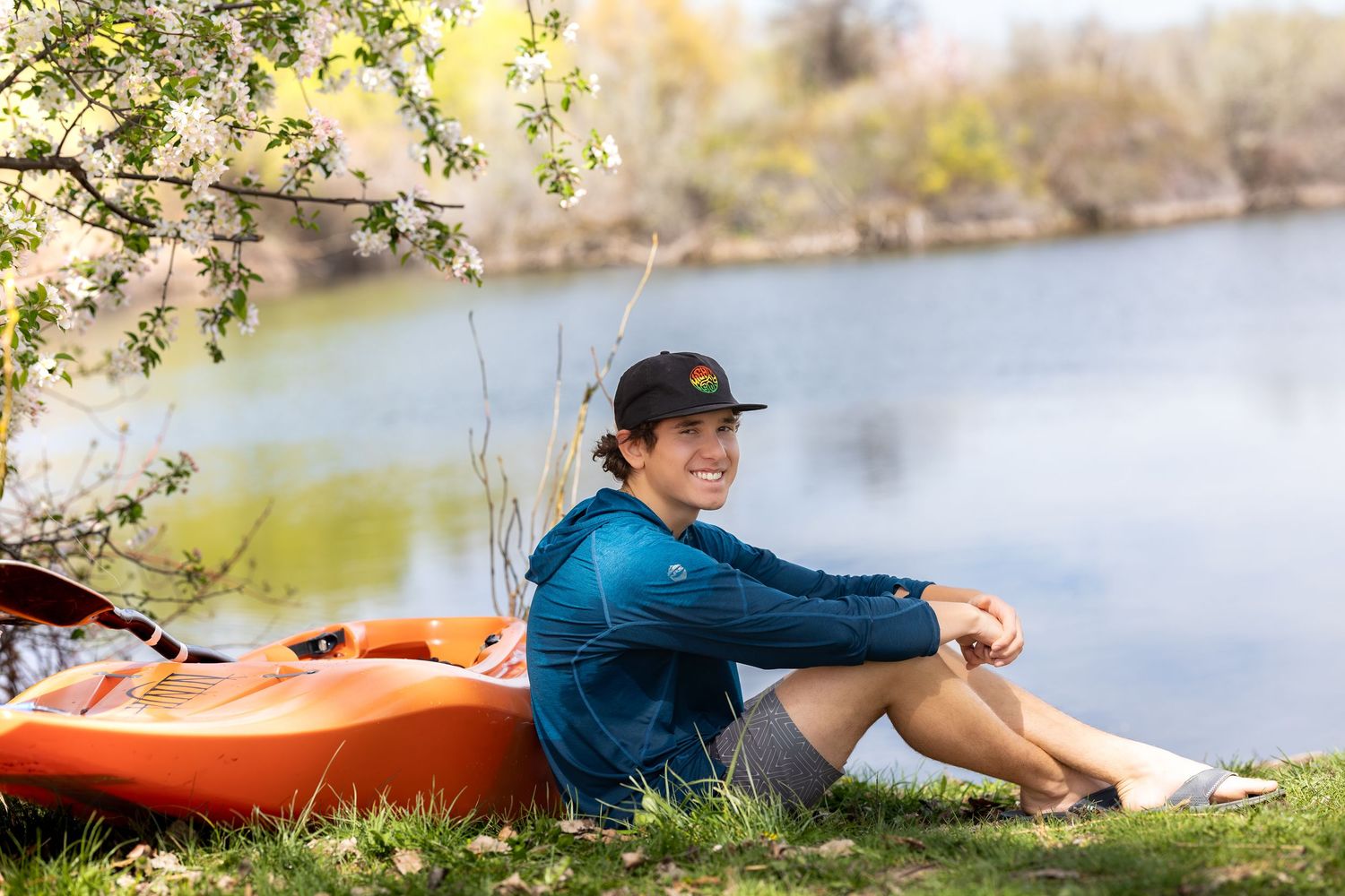 Smiling young man in blue shirt and cap sitting by a river with an orange kayak on a sunny day.