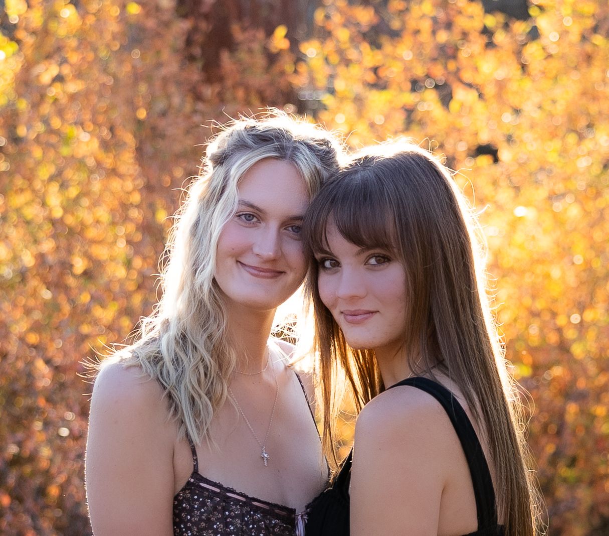 Two people posing together outdoors with autumn foliage creating a warm golden backdrop in sunlight.