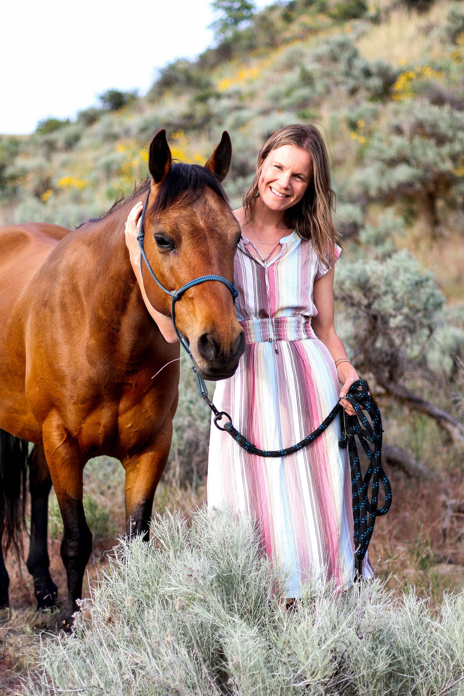 A bay horse with a black bridle stands next to someone in a striped dress against a sage brush hillside.