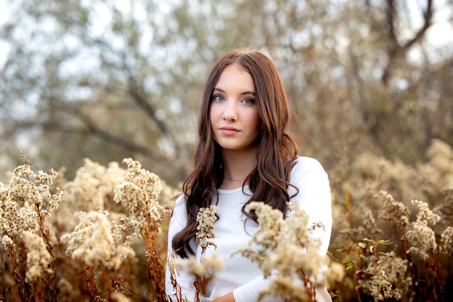 A person with long brown hair poses outdoors among tall dried grass and flowers in warm autumn sunlight.