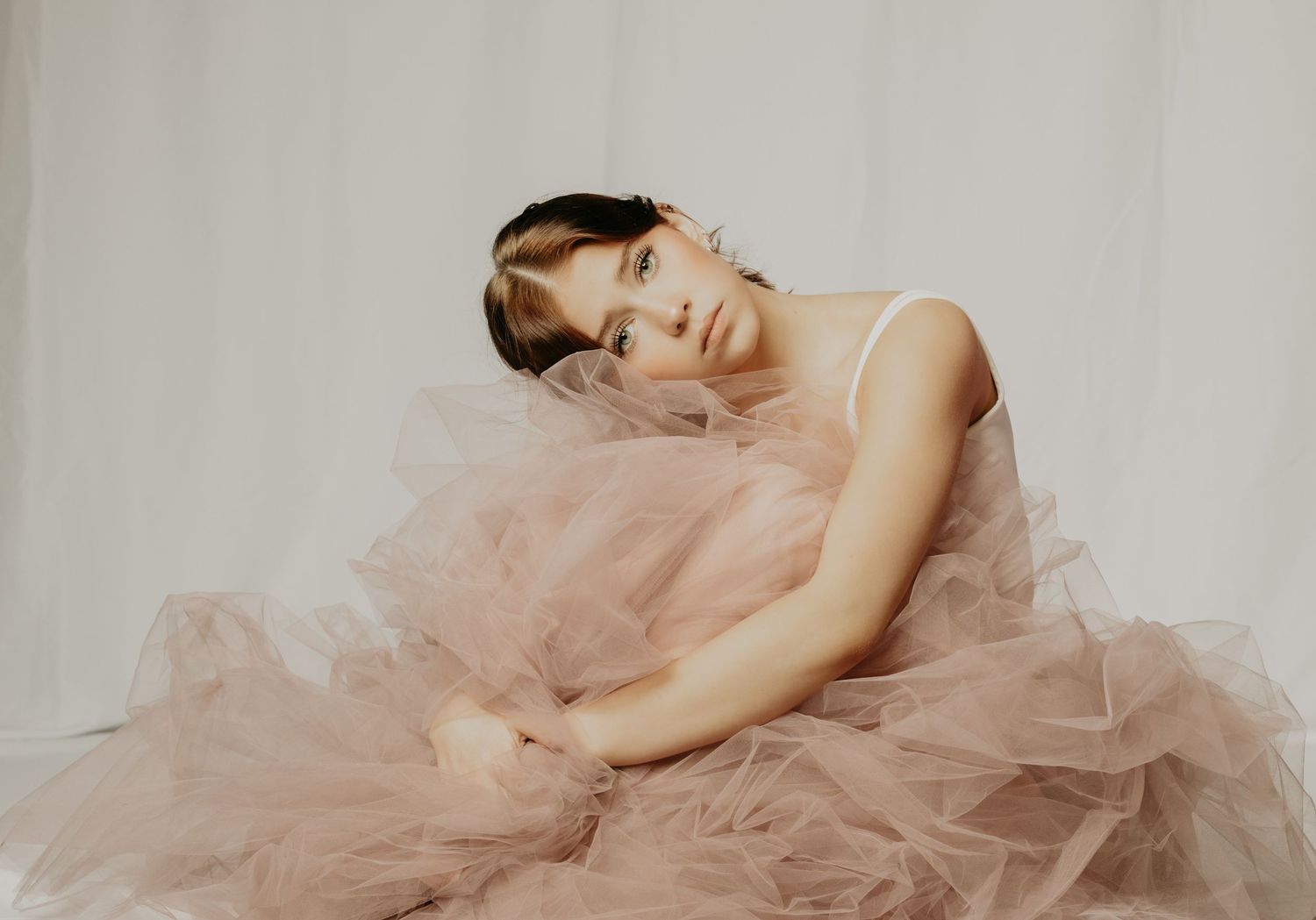 A graceful figure in a blush pink tulle gown poses elegantly against a light backdrop in a soft, dreamy portrait.