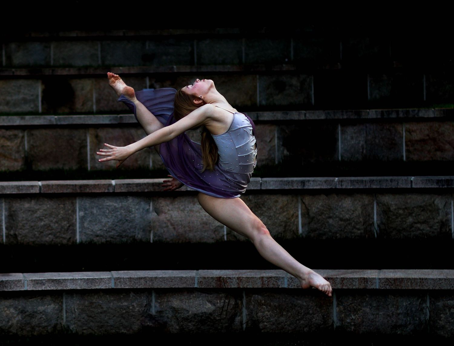 Dramatic dance leap captured on concrete steps in low light conditions.