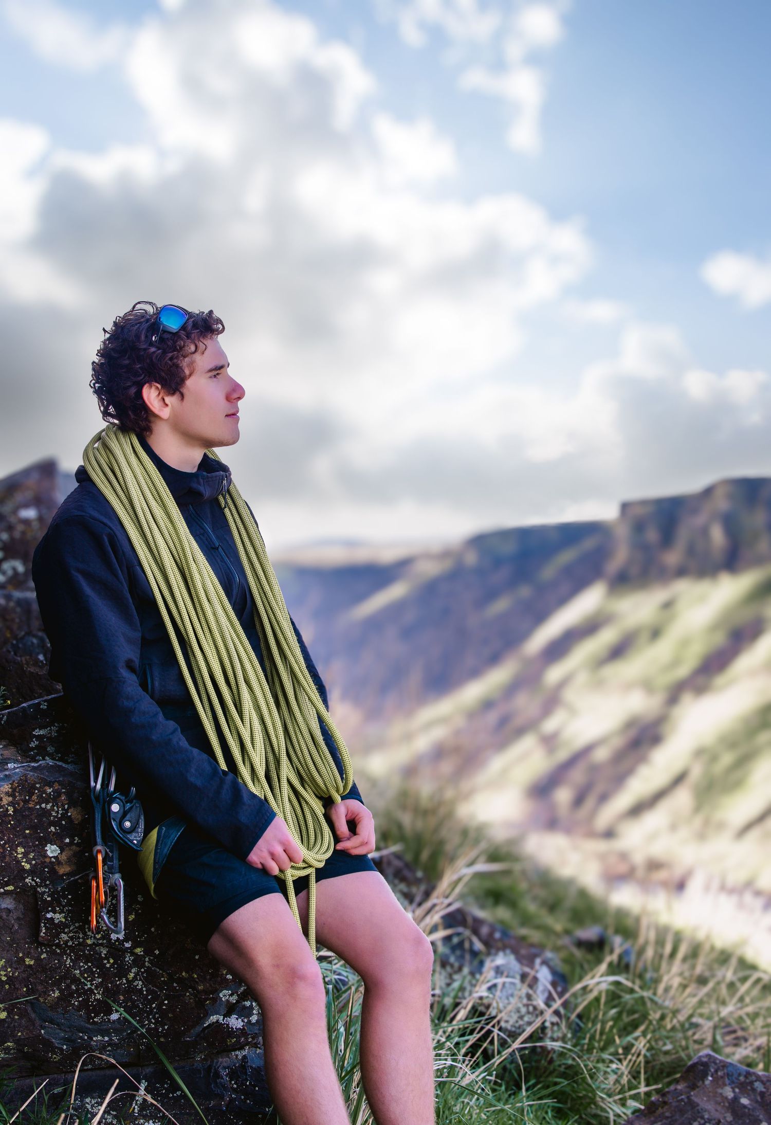 A person in a dark jacket and yellow scarf sits on a grassy hillside overlooking a mountainous coastal landscape.