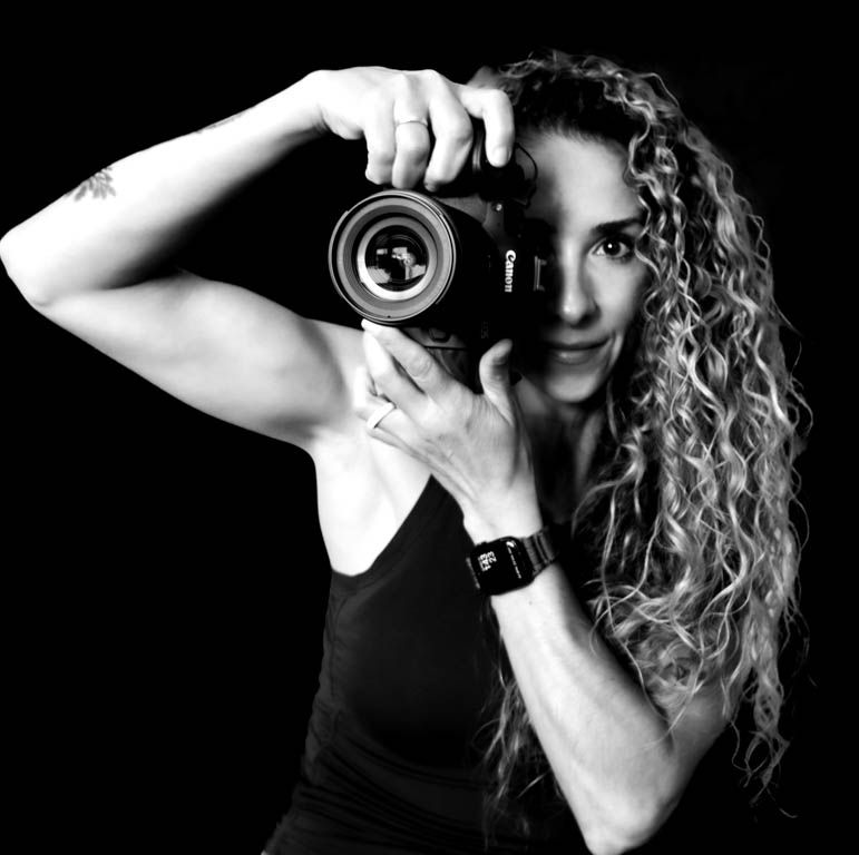 Black and white portrait of a photographer with curly hair holding a camera lens to their eye.