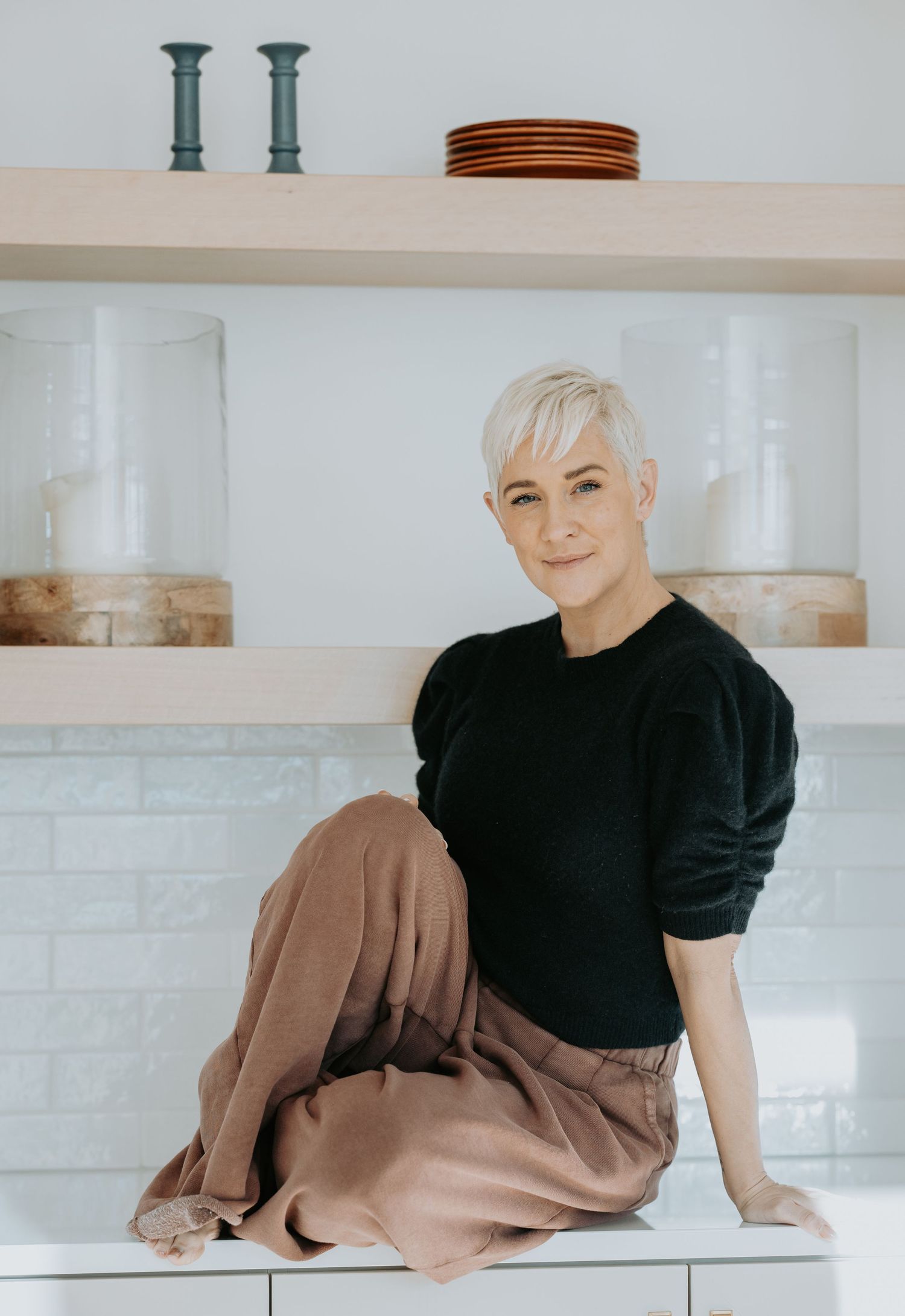 A person in a black top and beige pants sits casually on a white counter against a light-colored wall.