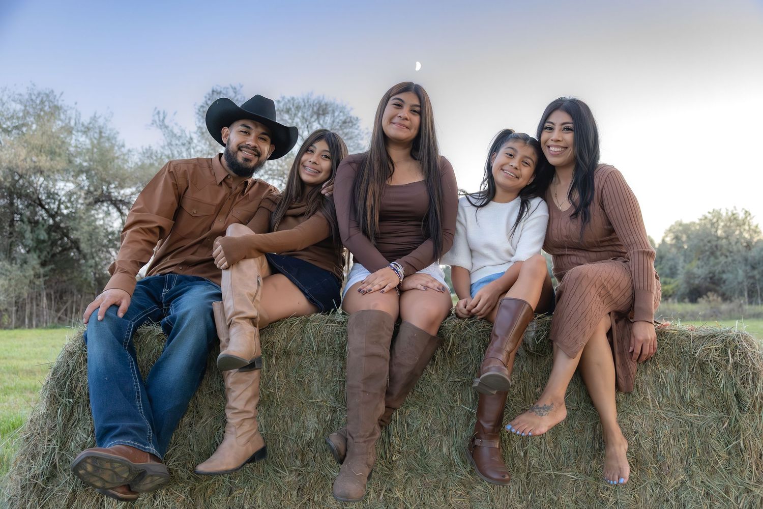 A family in coordinating brown outfits sits together on a hay bale outdoors in a rural setting during golden hour.
