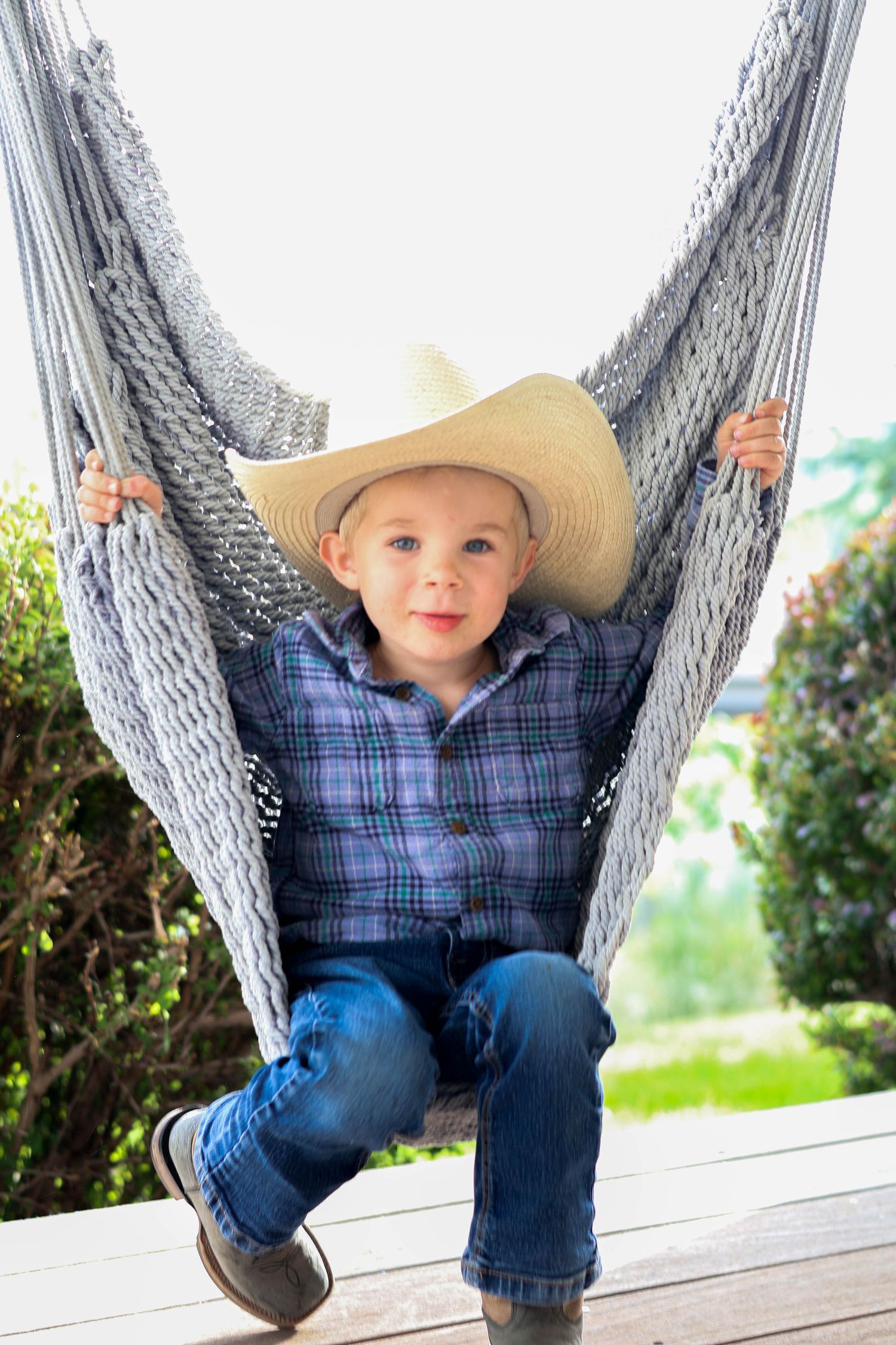 A young child wearing a cowboy hat and plaid shirt sits in a gray rope hammock.