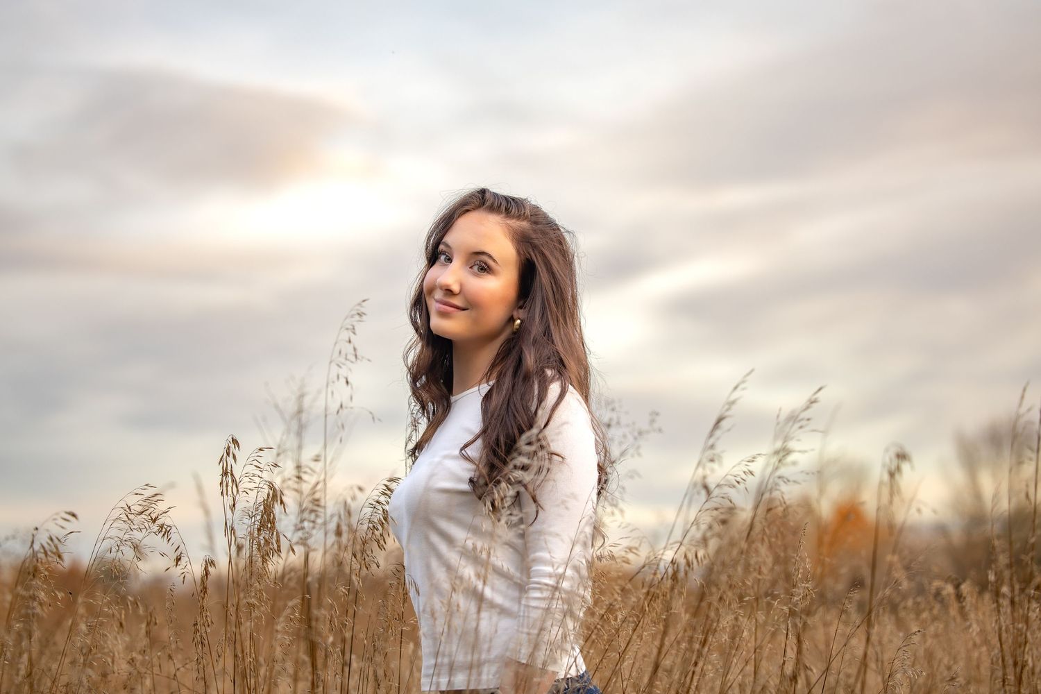 A woman in a white blouse stands thoughtfully in a golden wheat field at sunset.