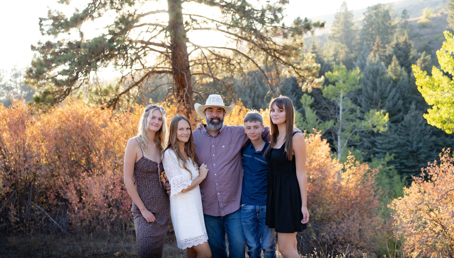 A group of people pose together outdoors during golden hour with mountains and trees in the background.