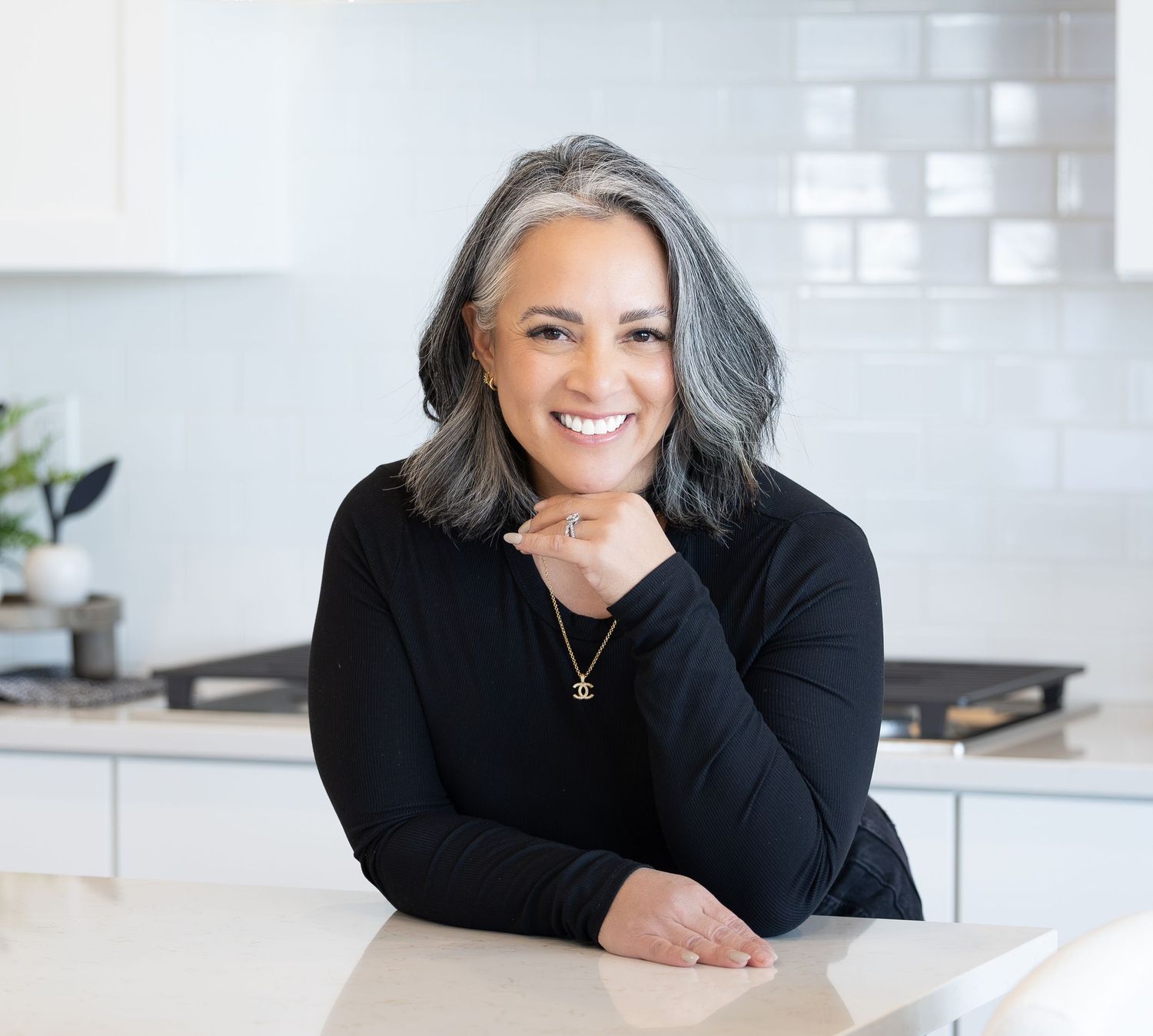 A person with silver gray hair wearing a black sweater smiles while resting their chin on their hand in a modern kitchen.