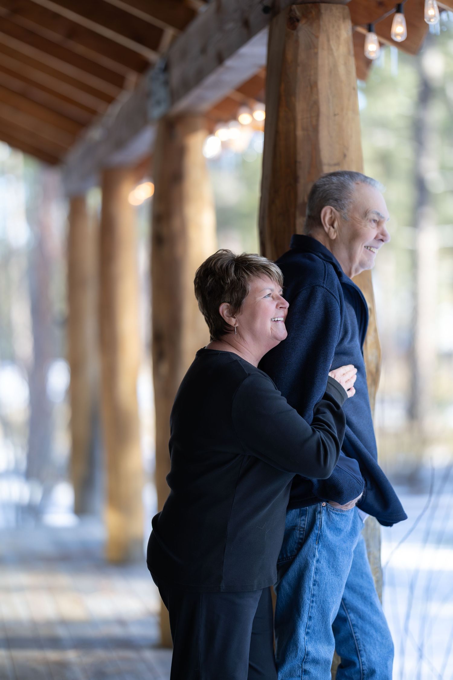A couple embraces in a tender moment under a wooden covered walkway with string lights overhead.