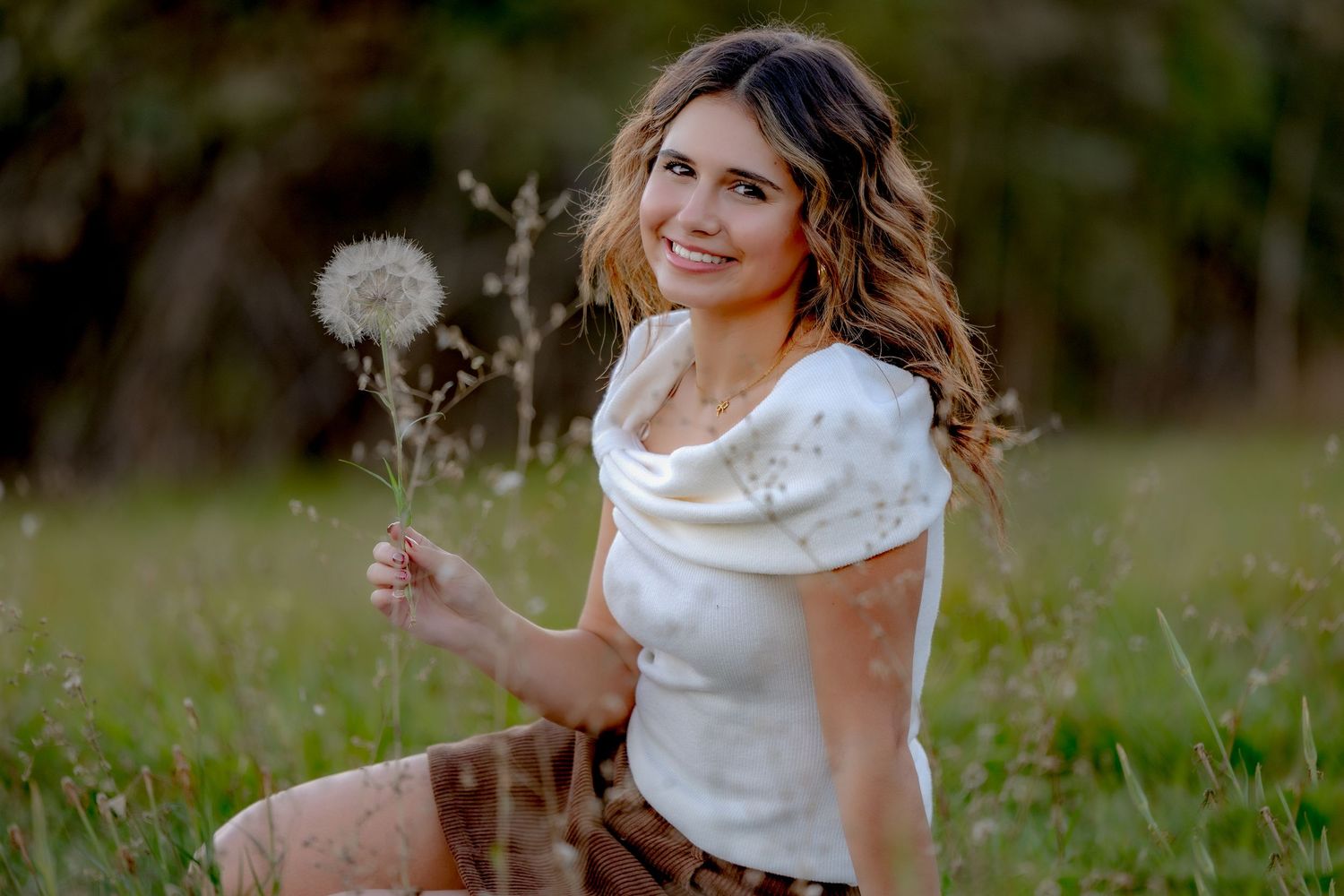 Someone in a white dress stands in a grassy field holding a dandelion flower during golden hour.