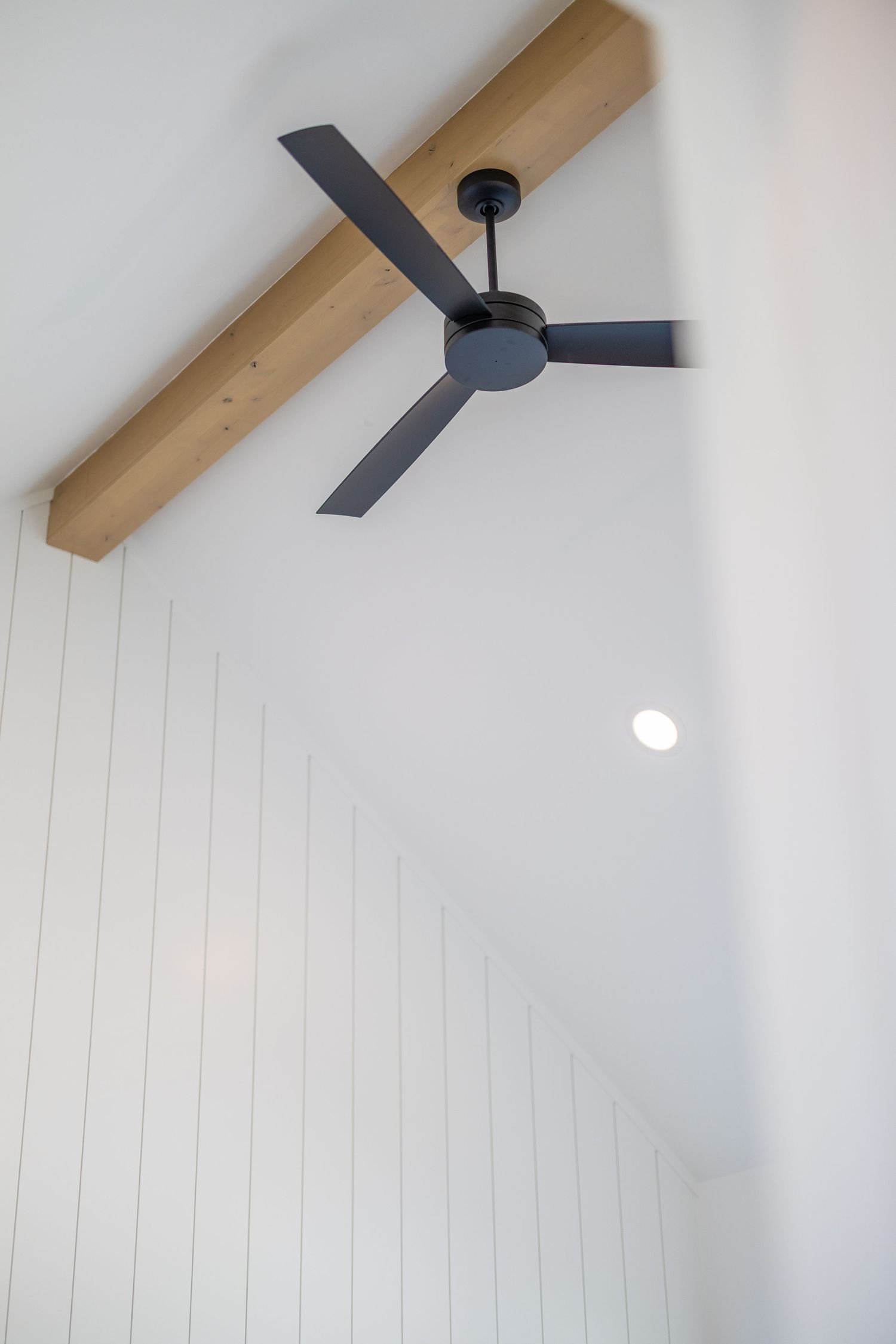 Modern black 3-blade ceiling fan mounted on vaulted white shiplap ceiling with exposed natural wood beam accent.