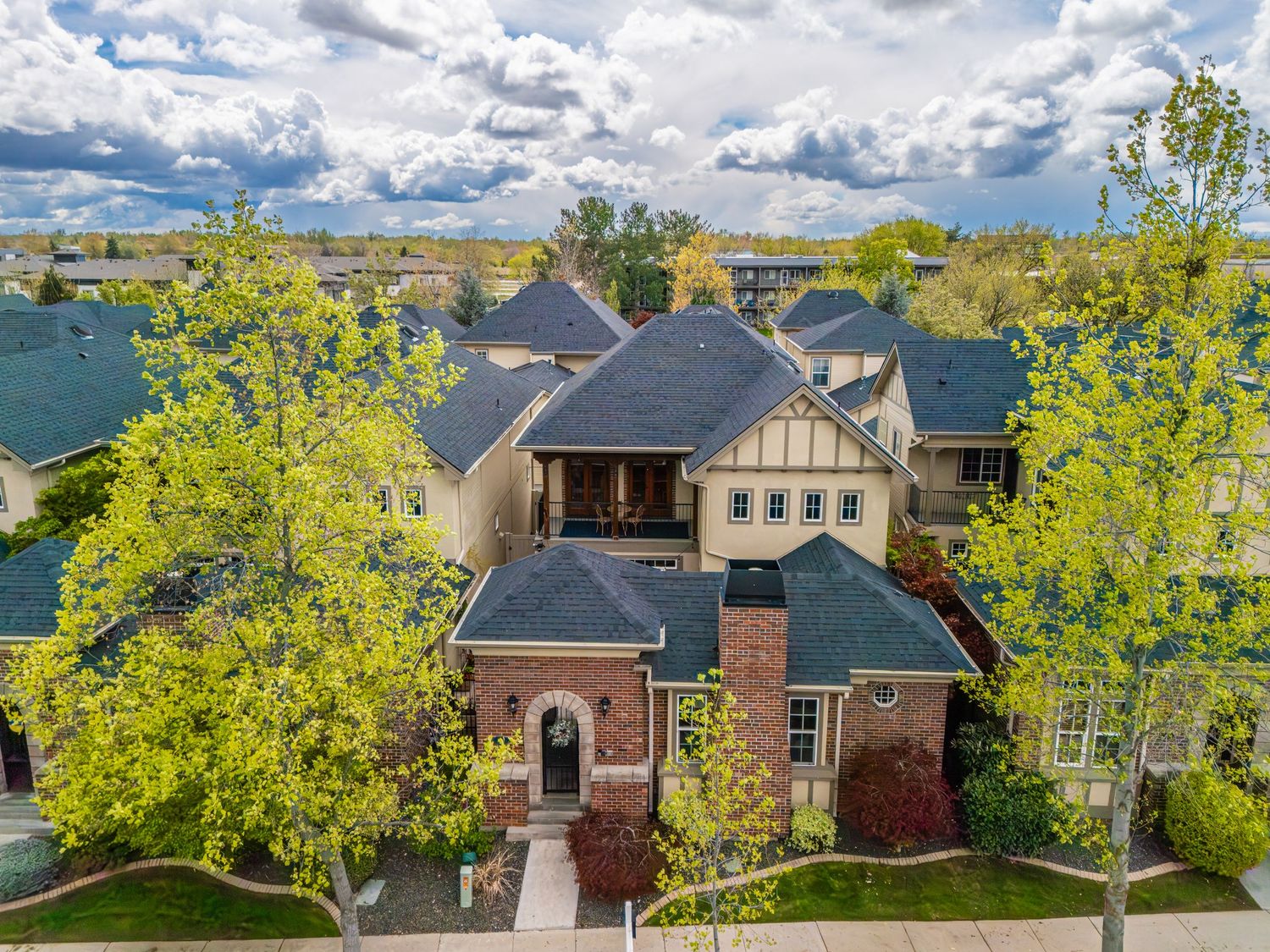 Aerial view of a brick and Tudor-style home surrounded by mature trees in a suburban neighborhood under cloudy skies.