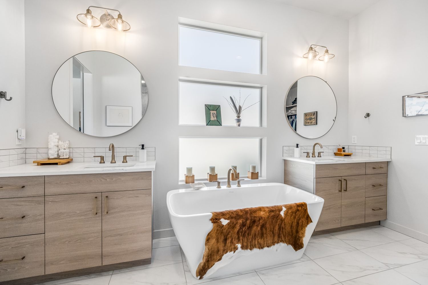 Modern master bathroom with freestanding tub, wood vanities, round mirrors, and white marble floors.