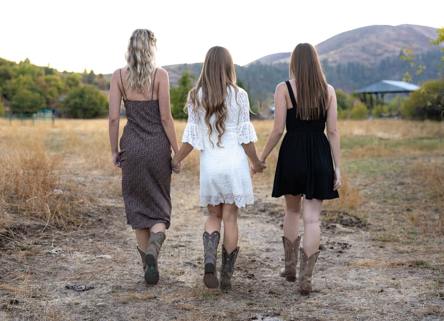 Three people walking away from camera on a dirt path through dry grass with mountains in the background.