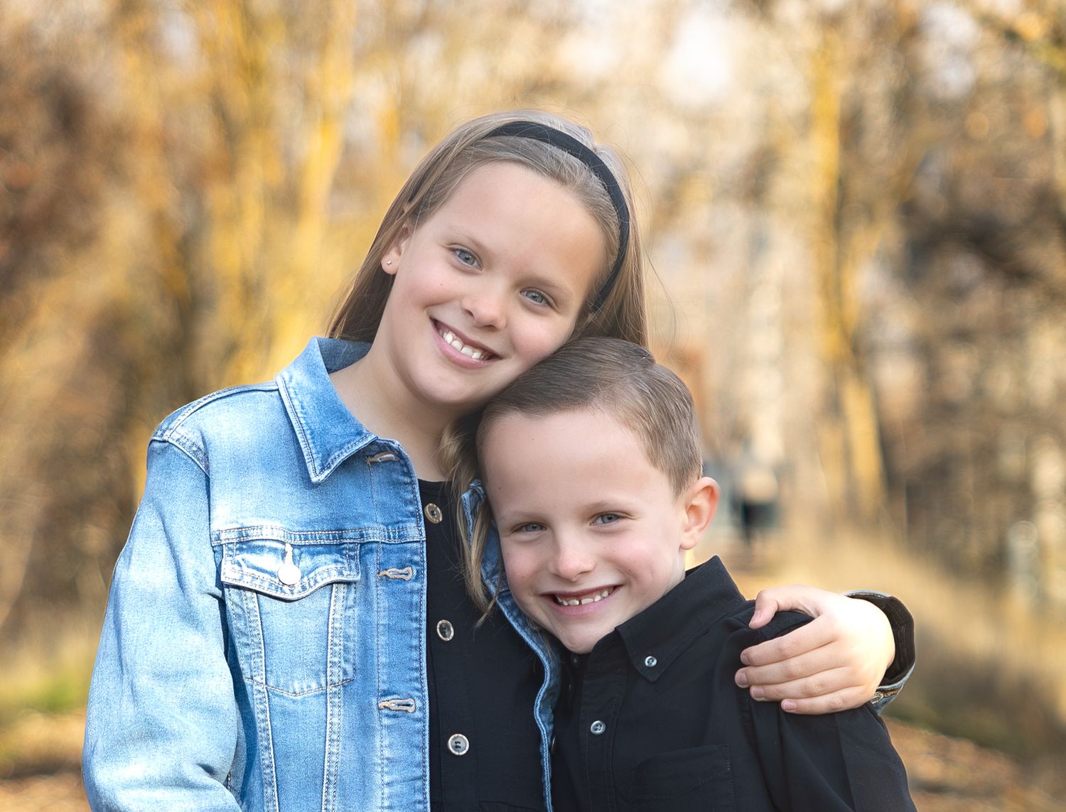 Children wearing denim and dark jackets pose playfully together outdoors in an autumn setting.