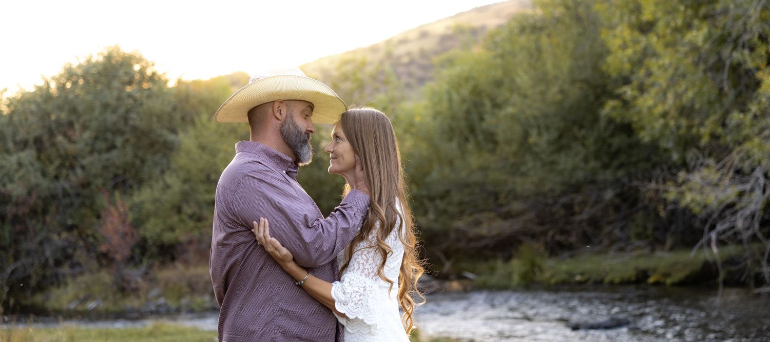 A couple embraces in a romantic outdoor photoshoot by a river with mountains and trees in the background.