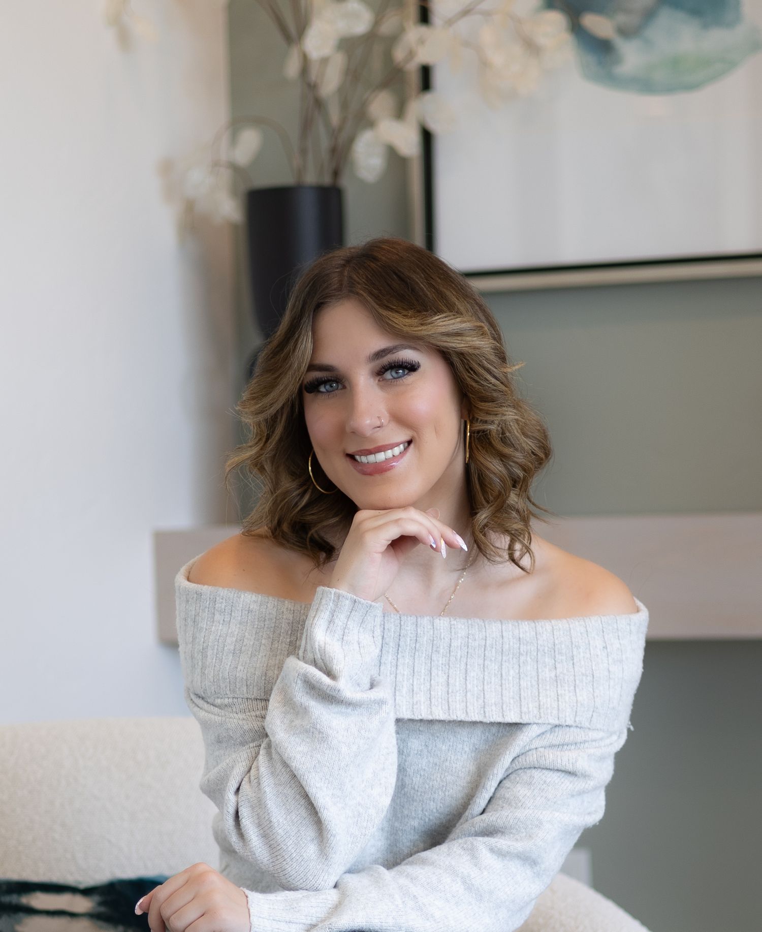 A person in a cozy off-shoulder grey sweater poses indoors while smiling warmly.
