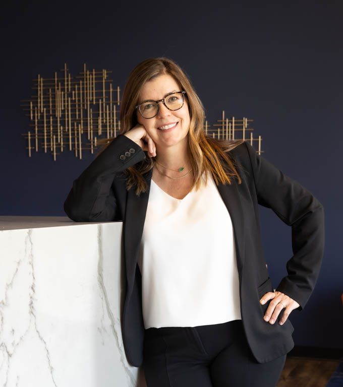 Professional business portrait with glasses and black blazer standing against dark wall with metallic wall decor.