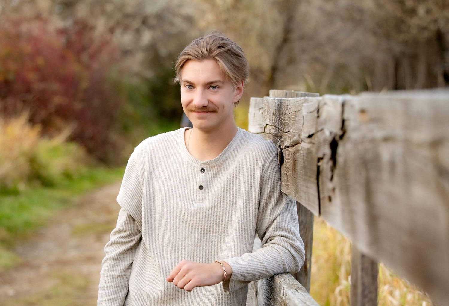 A person in a gray henley sweater stands against a stone wall in an outdoor setting during autumn.