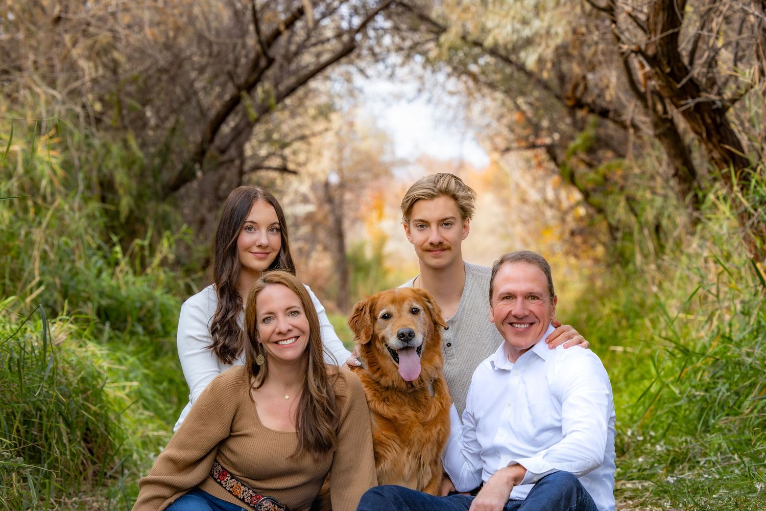 Family poses outdoors for autumn portraits with their golden retriever dog in a natural setting.