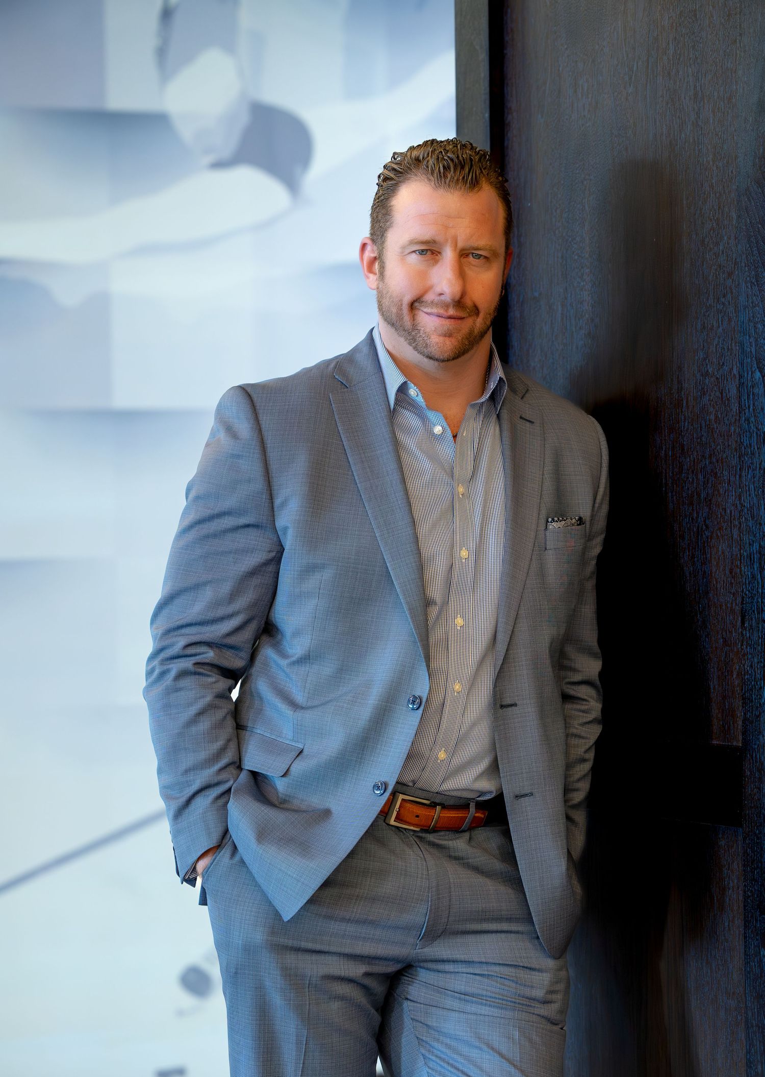 A man in a gray suit and striped dress shirt poses confidently against a wall in a professional portrait setting.