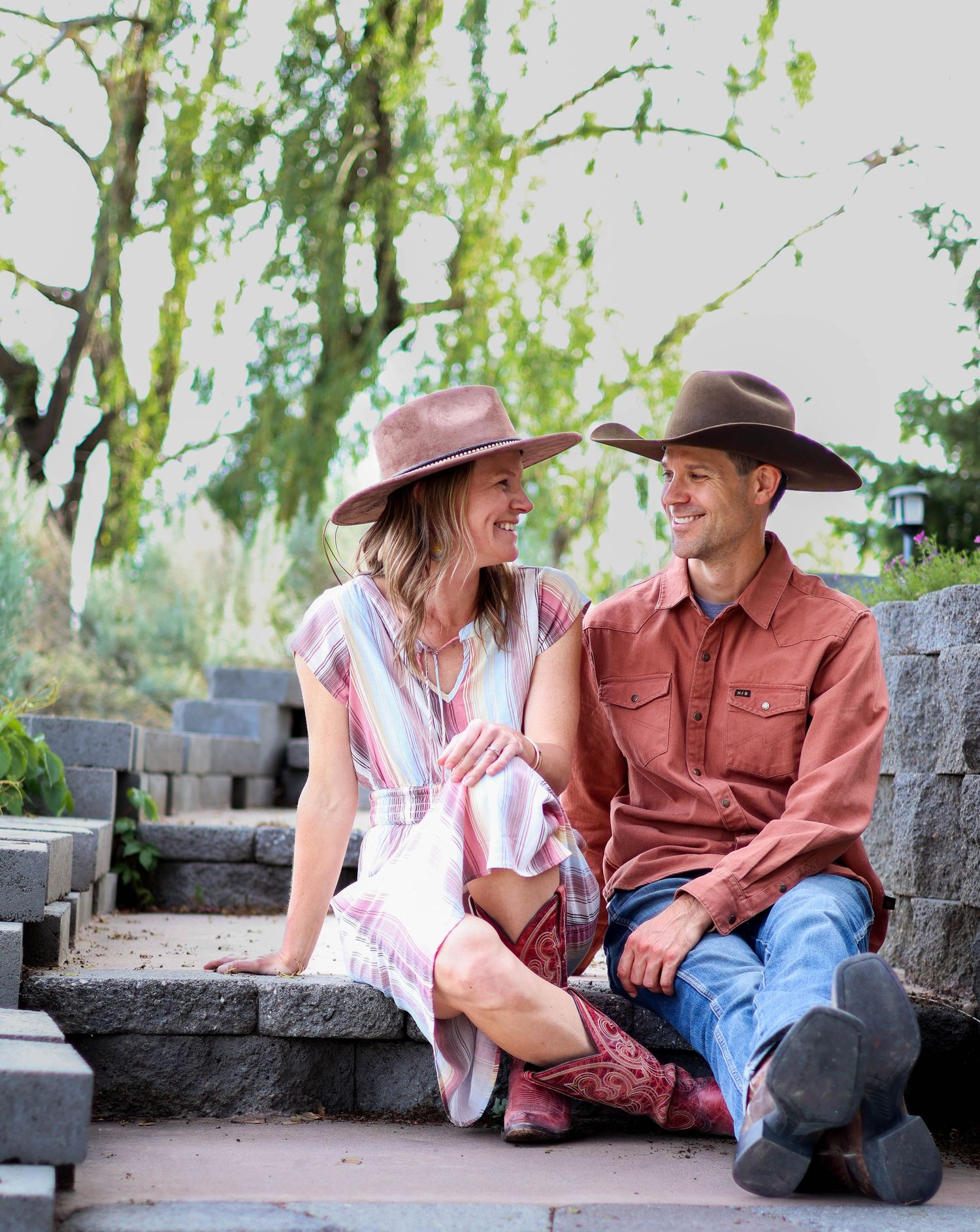 A couple in western attire sits together on stone steps under a weeping willow tree.