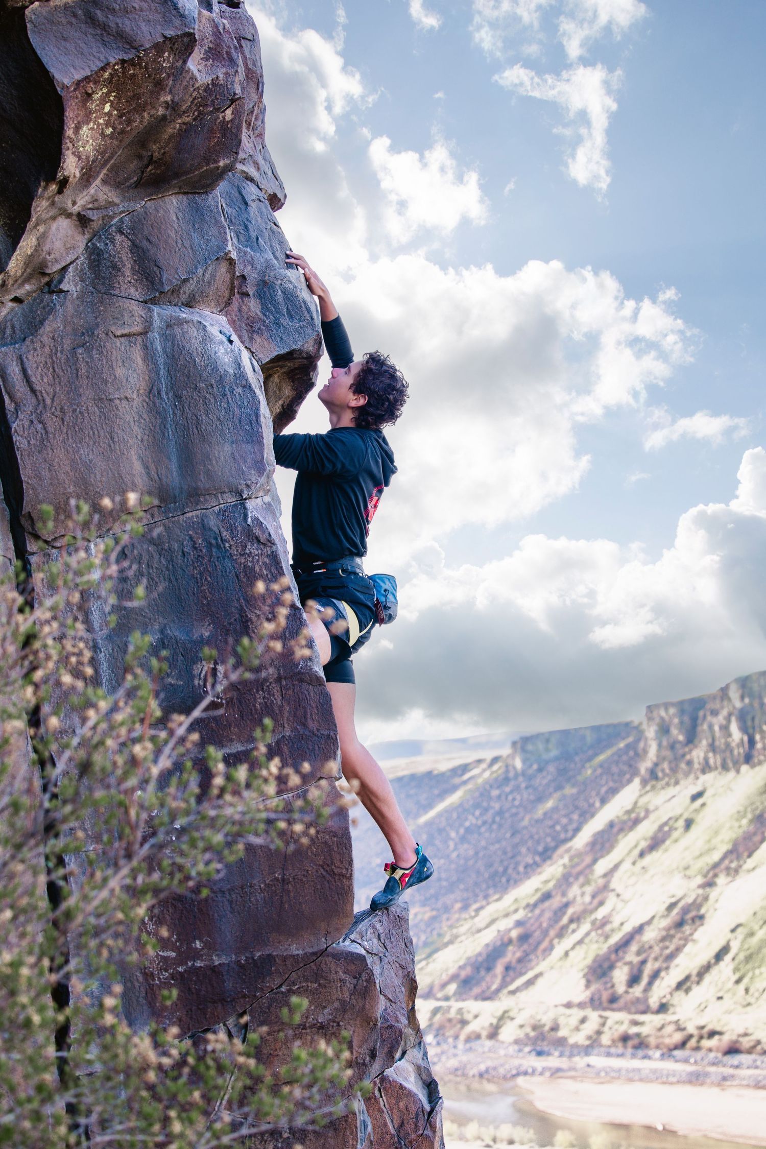 Rock climber scales steep cliff face with dramatic mountain valley landscape in background during outdoor bouldering session.
