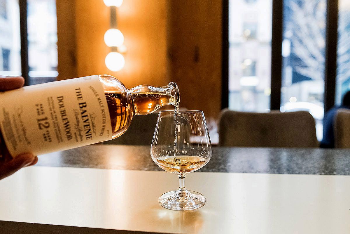 Amber whiskey being poured into a crystal snifter glass on a dark table with soft lighting and windows in background.