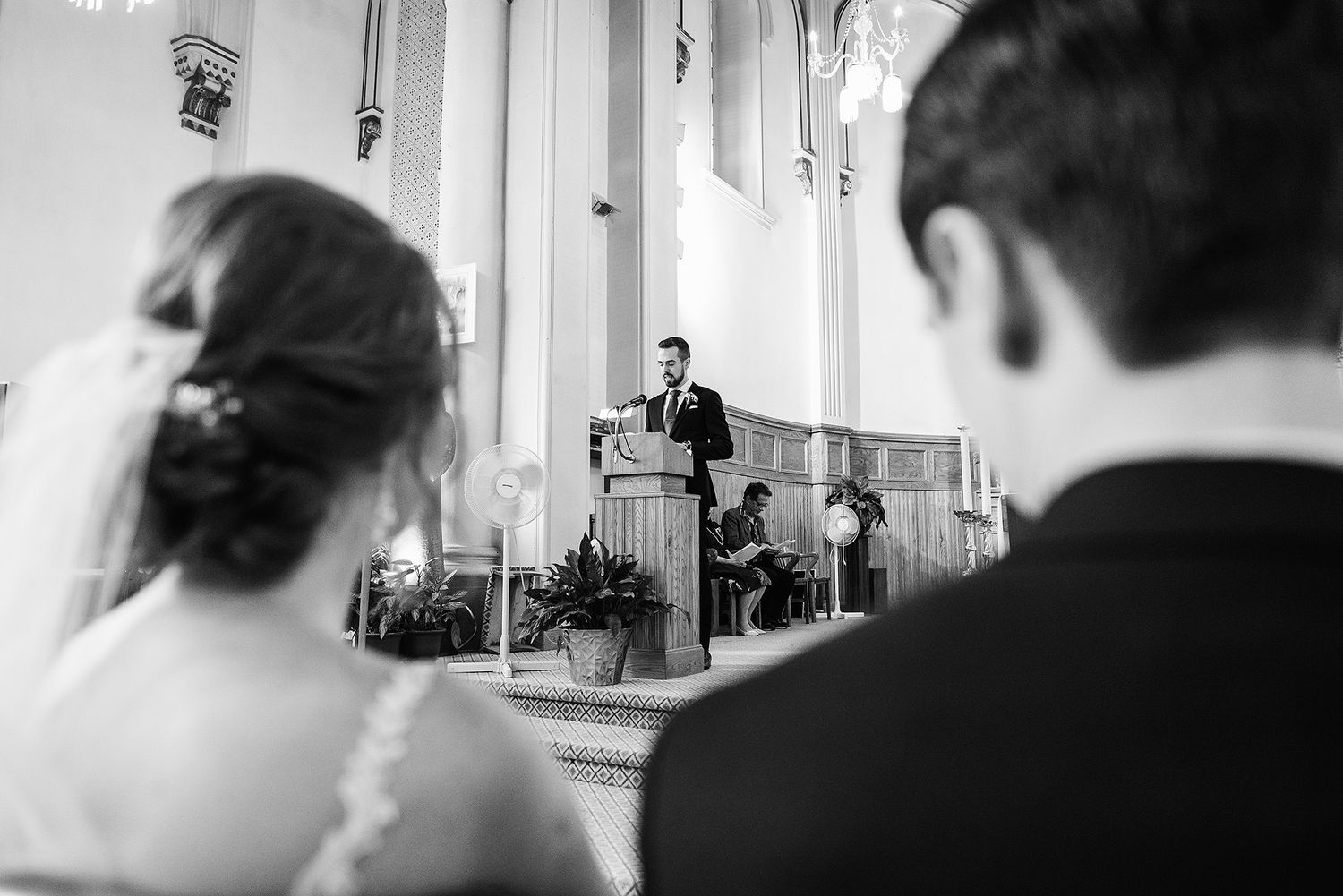 Black and white wedding ceremony photo shows minister speaking from pulpit while couple listens from front pew.