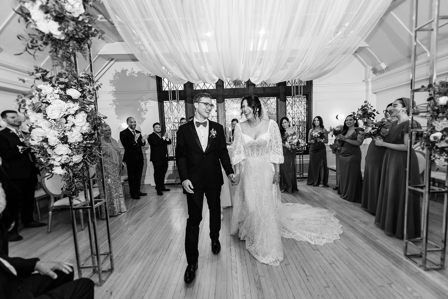 A black and white photo of a wedding couple walking down an elegantly draped aisle with guests watching from the sides. Club Atwater.