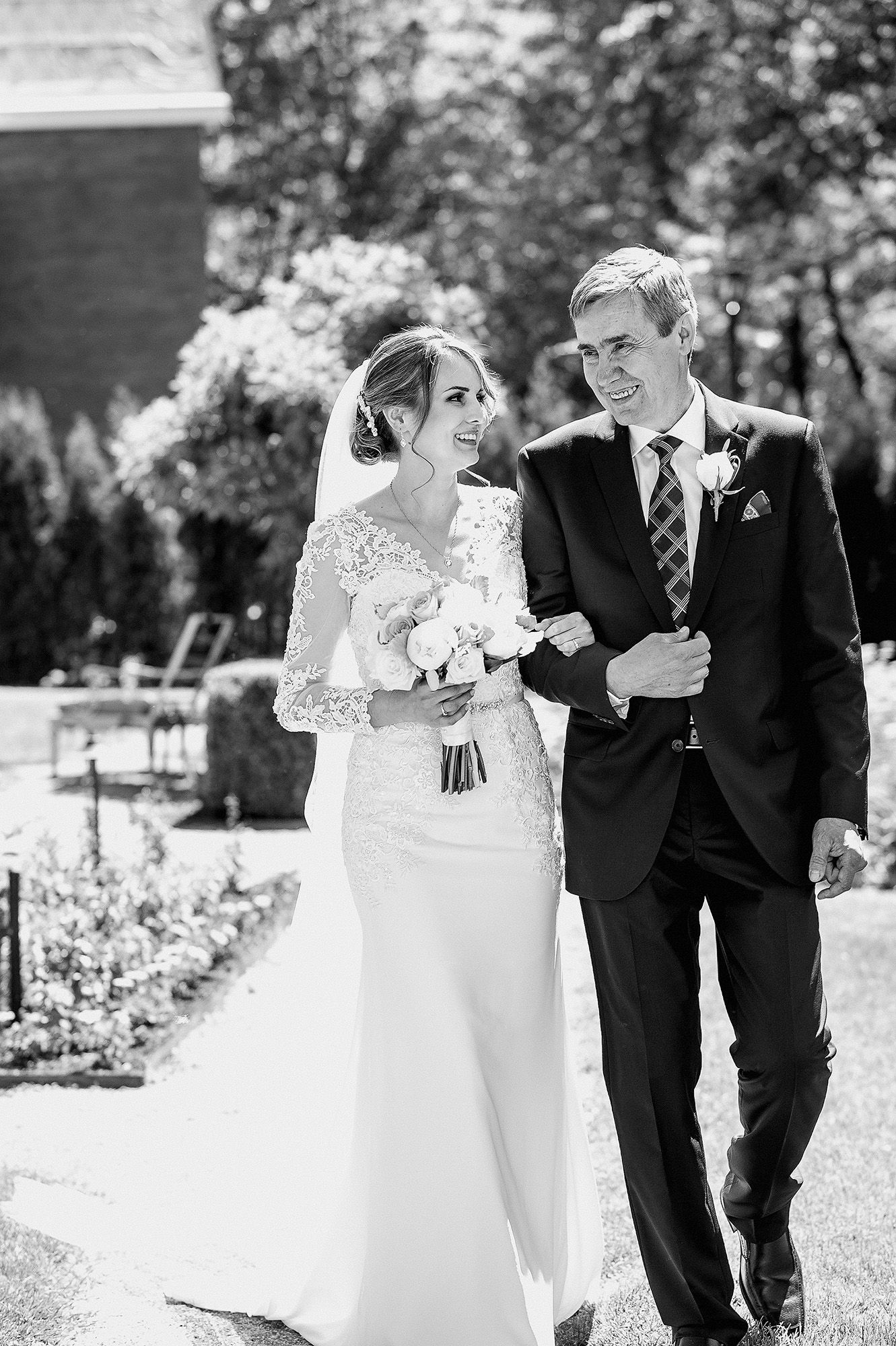 A joyful black and white wedding photograph capturing a couple walking outdoors in formal attire with lush greenery behind them.