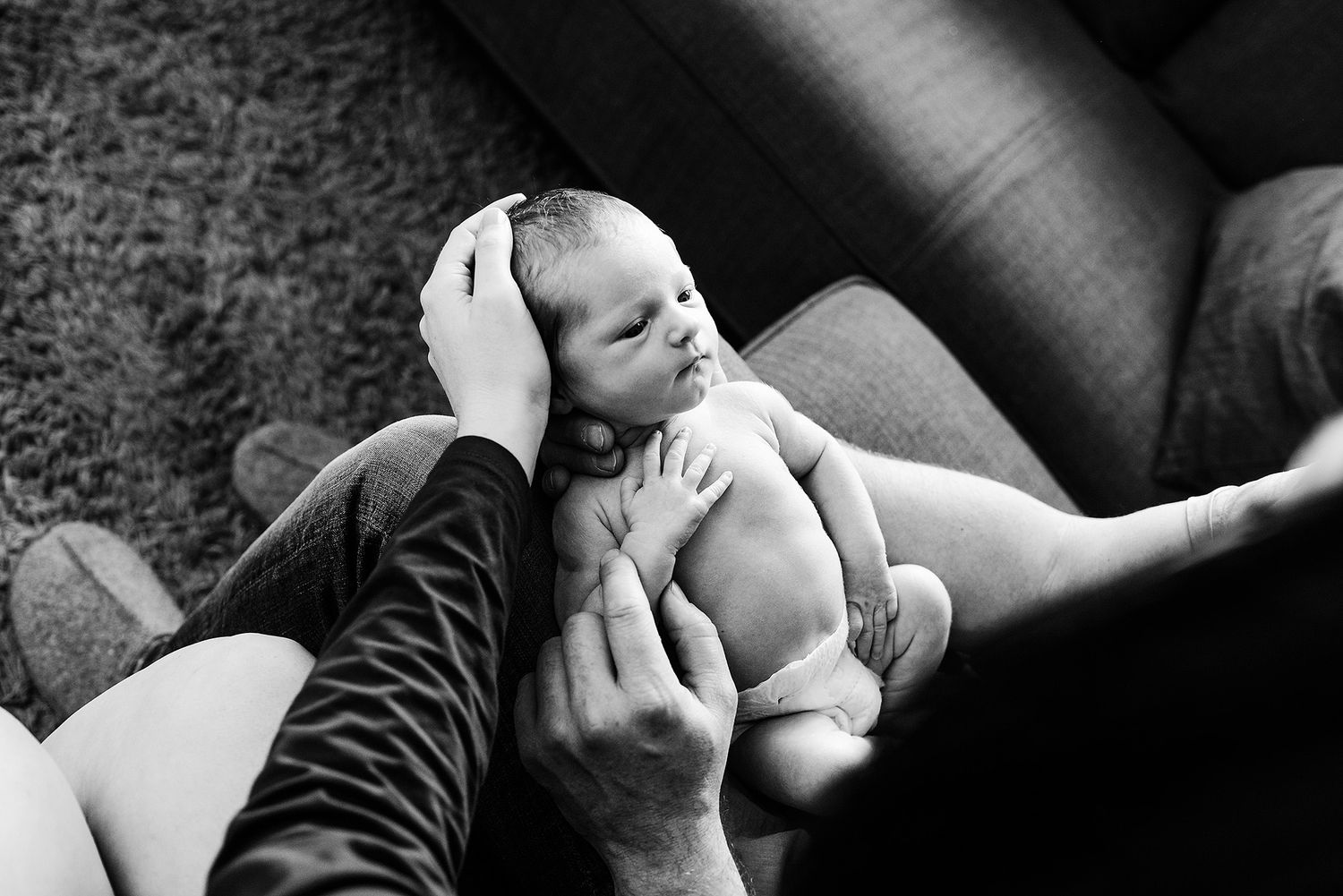 A tender black and white moment shows a young baby being cradled in someone's arms on a leather sofa.