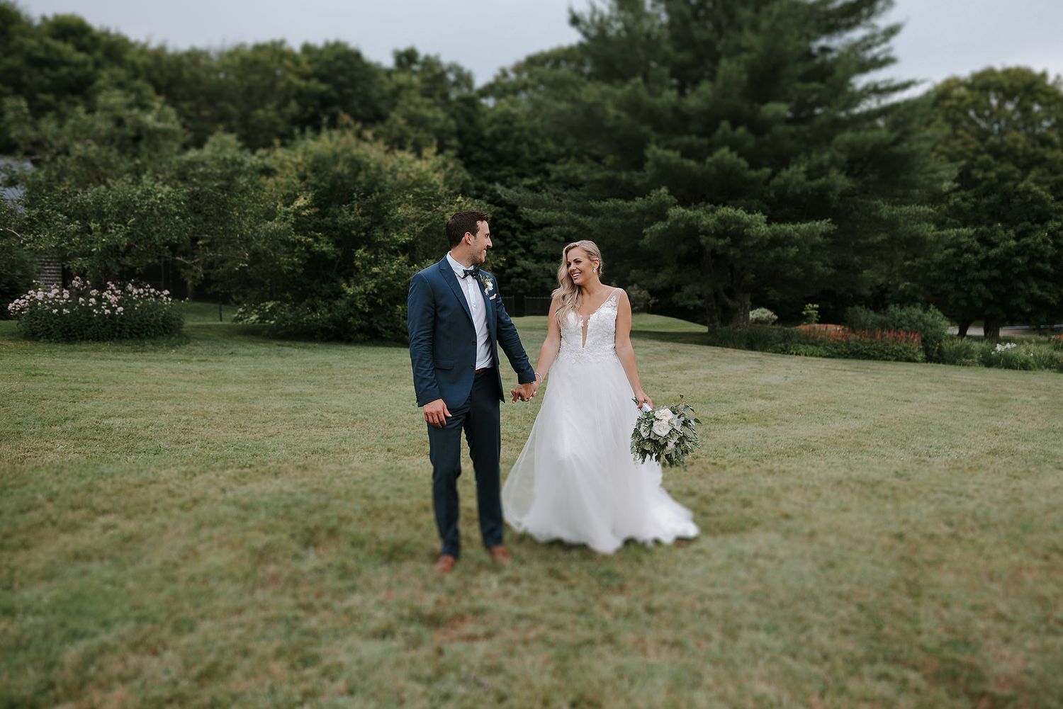 A couple in wedding attire holds hands while standing together on a lush green lawn surrounded by trees. Vignoble Le Ruisseau.