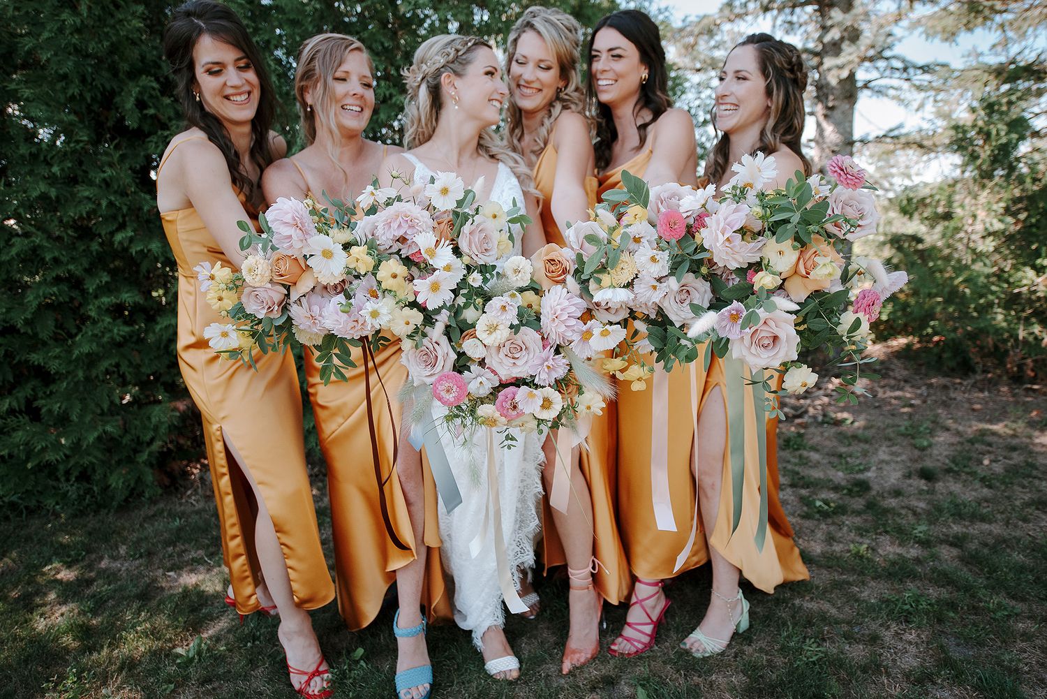 A group of bridesmaids in flowing yellow dresses holds pink and white bouquets while posing together outdoors.