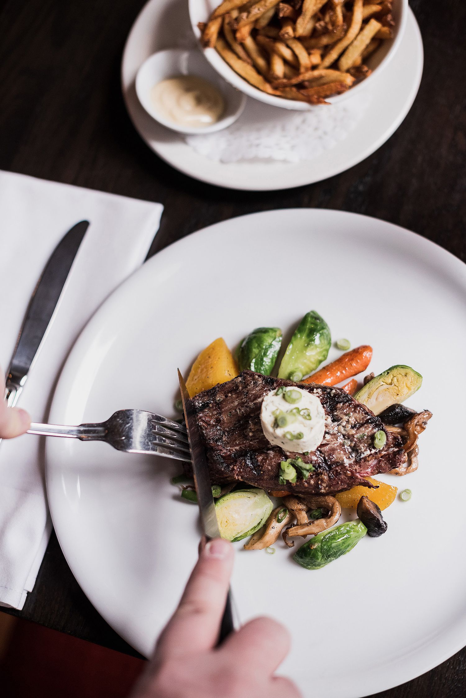 A grilled steak topped with herb butter served with roasted vegetables and a side of french fries on white plates.